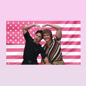 Könnte beinhalten: Ein rechteckiges Wandbanner mit einem rosa-weißen amerikanischen Flaggenmuster. Zwei Personen sind vor der Flagge positioniert und bilden beide mit ihren Armen über ihren Köpfen eine Herzform. Eine Person trägt ein schwarzes T-Shirt, die andere ein gestreiftes Hemd.