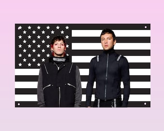 May include: A rectangular wall flag featuring a black and white American flag design with stars and stripes. Two individuals are positioned in the foreground, both wearing black jackets with white text accents against the monochromatic backdrop.