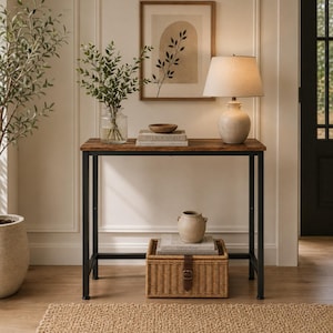 May include: A rectangular console table with a dark wood-grain top and a black metal frame. The table holds a beige lamp, a small wooden bowl, and two books titled Live Beautifully and This Is Home. A woven wicker storage basket sits below.