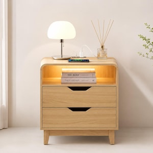 May include: A light wood nightstand with two drawers, an open top shelf with integrated lighting, and a charging pad on the surface. A white mushroom-shaped lamp, a glass reed diffuser, and a smartphone are placed on top of the nightstand.