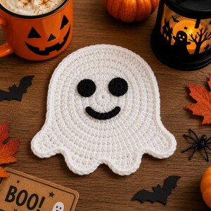 May include: A white crocheted ghost coaster featuring two round black eyes and a curved black mouth. The ghost shape has a rounded top and a scalloped bottom edge, crafted with a consistent stitch pattern to create a flat, circular-like surface.