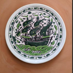 May include: A round white ceramic plate featuring a hand-painted dark green illustration of a sailing ship on the water. The design includes stylised waves, two small fish, and a decorative border of green leaves and dots encircling the centre.