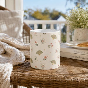 May include: A white ceramic mug featuring a repeating pattern of green folding fans, pink cherry blossom branches, and mahjong tiles with red and green characters. The design includes small gold star accents scattered across the surface.