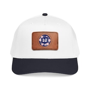 May include: A white baseball cap with a dark navy blue brim and a rectangular brown leather patch on the front. The patch features a circular design with pink flowers, green leaves, and the text BABY, surrounded by the phrase A baby is a blessing!