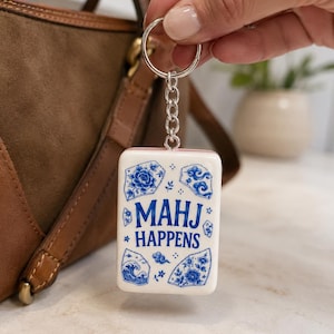 May include: A rectangular white keychain featuring blue decorative patterns and the text "MAHJ HAPPENS" in blue lettering. The design includes illustrations of flowers, clouds, and waves in a style reminiscent of blue and white porcelain.