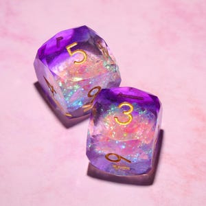 May include: Two translucent polyhedral dice featuring a deep purple gradient that transitions into a clear center filled with iridescent, shimmering flakes. Each die has engraved numbers filled with gold ink, displaying the digits five, nine, three, and six.