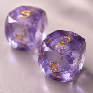 May include: Two translucent purple dice with faceted edges and gold-colored numbers. The die in the foreground displays the numbers two and six clearly, while the die in the background shows the number five on its top face.