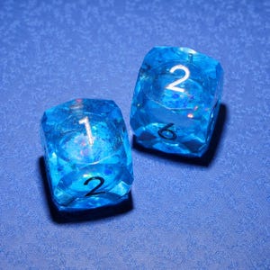 Könnte beinhalten: Zwei durchscheinende blaue sechsseitige Würfel mit einer facettierten, edelsteinartigen Oberfläche. Die Würfel enthalten eingebettete irisierende Glitzerpartikel. Die Zahlen sind in einer dunklen, fetten Schrift gedruckt, wobei die Zahlen 1, 2, 2 und 6 sichtbar sind.