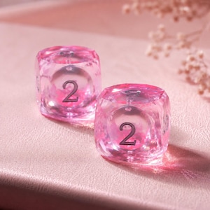 May include: Two translucent pink dice with a faceted, crystal-like texture. Each die features a prominent dark numeral 2 centered on the front face. The material appears clear and glossy, catching the light to create soft highlights and shadows.