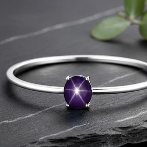 Könnte beinhalten: Ein dünner Silberarmreif mit einem einzelnen ovalen, tiefvioletten Sternsaphir in einer Vier-Krappen-Fassung. Der Edelstein zeigt einen ausgeprägten sechsstrahligen Sterneffekt, der sich in der Mitte seiner polierten, glatten Oberfläche befindet.