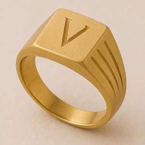 May include: A gold-toned signet ring with a brushed matte finish, featuring a square face engraved with the capital letter V and three parallel diagonal grooves etched into the side of the band, creating a textured and structured appearance.