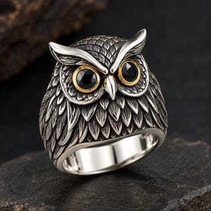 May include: A silver ring shaped like an owl head with detailed feather engravings. The owl features two prominent black circular eyes set within gold-toned rims, a sharp silver beak, and pointed ear tufts, creating a realistic bird appearance.