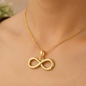 Pode incluir: Um colar de tom dourado com um pingente polido em forma de símbolo do infinito, suspenso por uma delicada corrente de elos. O pingente está centralizado e preso por uma pequena argola lisa, exibindo um design de laço contínuo e brilhante.