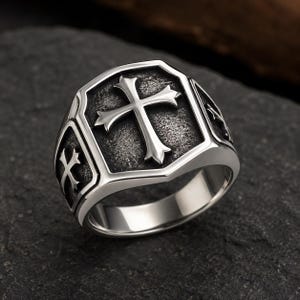 Peut inclure: Une bague en métal argenté présentant une face en forme de bouclier avec un emblème de croix en relief. Le fond du bouclier possède une finition oxydée, sombre et texturée. Des motifs de croix plus petits sont gravés sur les côtés de l'anneau.