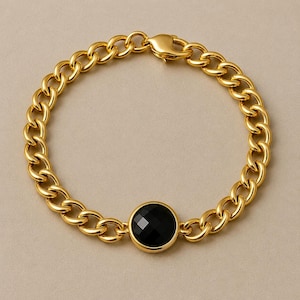 Puede incluir: Una pulsera de eslabones dorados que presenta una piedra negra redonda y facetada engastada en un bisel dorado pulido. La pulsera está construida con eslabones ovalados uniformes e incluye un cierre de mosquetón seguro en la parte superior.