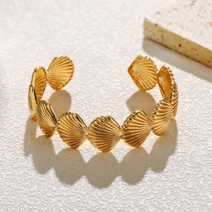 Peut inclure: Un bracelet manchette doré composé d'une série de motifs en forme de coquillages en éventail reliés entre eux. La surface métallique présente une finition polie et réfléchissante avec des stries rayonnantes imitant la texture des coquilles.