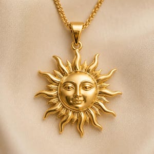 Peut inclure: Un collier pendentif en métal doré représentant un visage de soleil circulaire avec des rayons ondulés rayonnants. Le soleil présente un visage humain détaillé avec des yeux en amande, un nez et un léger sourire. Le pendentif est suspendu à une chaîne dorée.