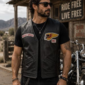 May include: A black leather motorcycle vest featuring a Hells Angels patch on the right chest and a winged skull emblem with an MC patch on the left chest. The vest includes side lacing details, two front pockets, and a smooth, matte finish.