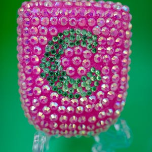 Può includere: Un adattatore per presa a muro rettangolare completamente ricoperto di strass rosa iridescenti. Al centro presenta una lettera C prominente formata da strass più piccoli di colore verde scuro. L'adattatore ha due poli metallici sulla parte superiore.