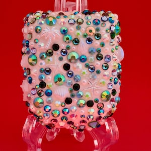 May include: A rectangular light pink object decorated with a dense arrangement of multicolored iridescent rhinestones, small white pearls, and embossed coastal motifs including starfish, seashells, and palm trees, supported by a clear stand.