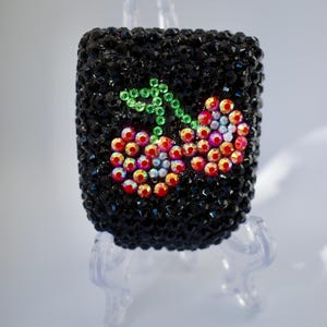 May include: A rectangular case covered in black faceted rhinestones featuring a central cherry design made of iridescent red and clear crystals with green crystal stems. The object is displayed on a clear plastic stand against a plain background.