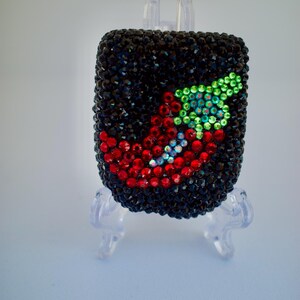 Può includere: Un oggetto rettangolare ricoperto di strass neri con un disegno di peperoncino realizzato con strass rossi e verdi. Il peperoncino ha un corpo rosso curvo e un gambo verde, con accenti iridescenti integrati nel design del peperoncino.