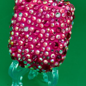 May include: A rectangular object covered in numerous pink and iridescent rhinestones of varying sizes, creating a dense, sparkling surface. The object is displayed on a translucent, light green plastic stand with curved legs and a central support.
