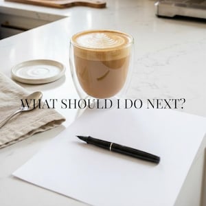 May include: A double-walled glass mug filled with a latte featuring leaf-patterned foam art, a small white ceramic saucer, a silver spoon, a black fountain pen, and a blank sheet of white paper with the text WHAT SHOULD I DO NEXT? printed in black.