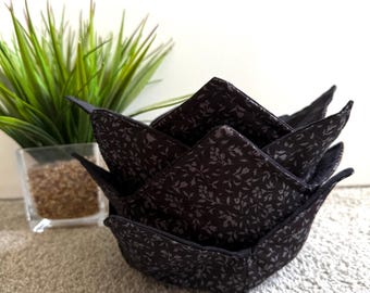 May include: A black fabric bowl cosy featuring a subtle, repeating dark grey floral pattern. The bowl cosy has a square-shaped base with four pointed, upward-facing corners, constructed from quilted material with visible stitching along the edges.