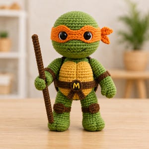 May include: A green crocheted turtle doll featuring a bright orange eye mask, a yellow plastron, and a brown belt with a yellow letter M. The figure has large black eyes and holds a long, thin, textured brown staff in its right hand.