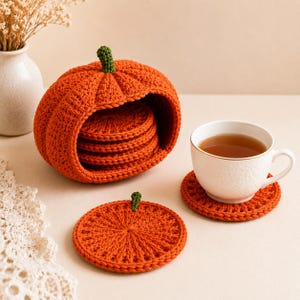 May include: A set of orange crochet pumpkin-shaped coasters with green stems. The coasters are stored inside a matching hollow pumpkin-shaped crochet holder. One coaster sits flat on the surface, and another supports a white ceramic teacup.