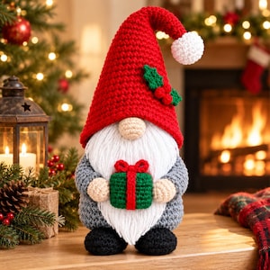 May include: A crocheted gnome figurine featuring a tall red hat with a white pom-pom and green holly leaves, a long white yarn beard, a beige nose, grey sleeves, black feet, and holding a small green gift box with a red ribbon and bow.