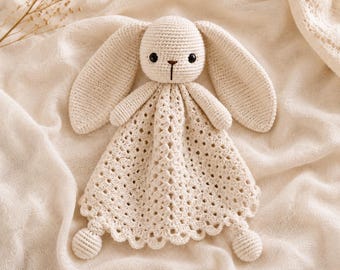 May include: A cream-colored crochet security blanket featuring a stuffed rabbit head with long ears, black bead eyes, and a brown stitched nose. The body consists of a triangular, open-work lace pattern with a scalloped edge and two small round feet.