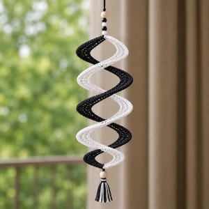 Könnte beinhalten: Ein hängendes Spiral-Windspiel aus schwarzem und weißem Garn mit einem Doppelhelix-Design. Die Struktur ist an einer schwarzen Kordel befestigt, die mit kleinen Holz- und schwarzen Perlen verziert ist und in einer passenden schwarz-weißen Quaste endet.