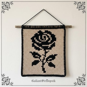 Op de afbeelding: Een rechthoekige gehaakte wandhanger met een zwart rozenontwerp op een beige achtergrond. Het stuk hangt aan een houten stok met een zwart koord. De tekst GallantSirDupsik is in het zwart onderaan de beige stof gedrukt.
