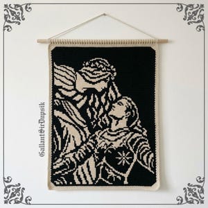Op de afbeelding: Een rechthoekige gehaakte wandhanger met een zwart en crèmekleurig grafisch ontwerp van twee figuren in een omhelzing. Het stuk hangt aan een houten stok met een touwtje. De tekst GallantSirDupsik staat verticaal aan de linkerkant gedrukt.