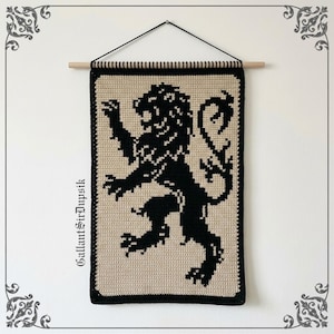 Puede incluir: Un tapiz rectangular de ganchillo que presenta la silueta de un león negro sobre un fondo beige. La pieza está suspendida de un palo de madera de color claro con un cordón negro. El texto GallantSirDupsik está impreso verticalmente a la izquierda.