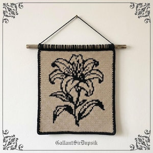 May include: A square crochet wall hanging featuring a black lily flower design on a beige background. The textile is suspended from a wooden dowel with a black cord for hanging. The text GallantSirDupsik is printed at the bottom of the image.