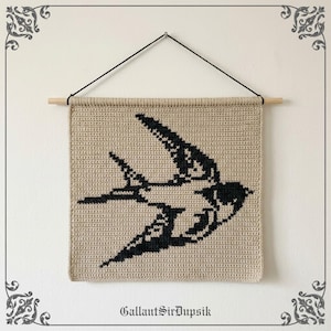 Op de afbeelding: Een vierkante gehaakte wandhanger met het silhouet van een zwarte zwaluw op een beige achtergrond. Het textiel hangt aan een lichte houten stok met een zwart koord. De tekst GallantSirDupsik staat onderaan de afbeelding.