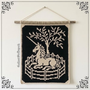 Puede incluir: Un tapiz rectangular de ganchillo que muestra un unicornio de color crema descansando dentro de una valla bajo un árbol sobre un fondo negro. La pieza cuelga de un palo de madera con una cuerda e incluye el texto GallantSirDupsik.