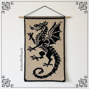 Op de afbeelding: Een rechthoekige gehaakte wandhanger met een zwart draken-silhouet op een beige achtergrond. Het werk hangt aan een lichtgekleurde houten stok met een zwart koord. De tekst GallantSirDupsik staat verticaal gedrukt aan de linkerzijde.