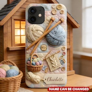 May include: A phone case with a quilted pastel background featuring 3D-style yarn balls, wooden knitting needles, a basket of yarn, a knitted swatch, buttons, and a rectangular tag with the name Charlotte printed in a script font.