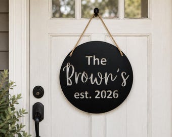 Pode incluir: Uma placa circular preta pendurada por uma corda de juta, com o texto branco The Brown's e est. 2026. A placa é feita de um material plano com letras recortadas, exibindo um design limpo e de alto contraste para uma porta.