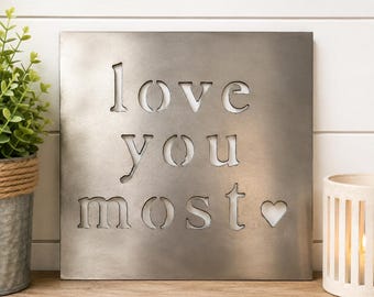 Pode incluir: Uma placa de parede quadrada de metal prateado escovado com o texto recortado "love you most" seguido por um pequeno símbolo de coração. As letras são em estilo estêncil, permitindo que a luz passe pelas aberturas na superfície metálica.