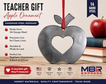 Pode incluir: Um ornamento cinza em forma de maçã feito de aço de calibre 14 com um recorte em forma de coração no centro. O ornamento está pendurado por uma fita vermelha. O texto na imagem inclui TEACHER GIFT, Apple Ornament, HANDMADE STEEL KEEPSAKE e 14 GAUGE STEEL.