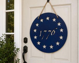 Pode incluir: Uma placa de parede circular de madeira azul-marinho com o número 1776 recortado no centro, rodeado por um anel de treze estrelas recortadas. A placa está suspensa por uma corda de juta bege presa a um gancho preto numa porta branca.
