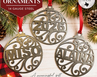 Pode incluir: Três ornamentos circulares de aço de calibre 14 com recortes intrincados de volutas. Um ornamento apresenta o ano 2026, enquanto os outros são personalizados com os nomes MISO e ALAINA. Cada peça é suspensa por uma fita vermelha.