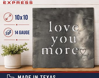 Pode incluir: Uma placa de parede quadrada de metal de 25,4x25,4 cm e calibre 14 com um acabamento escuro e escovado. O texto "love you more" está recortado no centro, seguido por um pequeno símbolo de coração. A placa é exibida ao lado de uma planta verde.