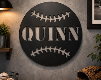 Pode incluir: Uma placa de parede circular de metal preto em forma de bola de beisebol, apresentando o nome QUINN recortado em letras maiúsculas grandes no centro, ladeado por duas fileiras horizontais de padrões de costura de beisebol.