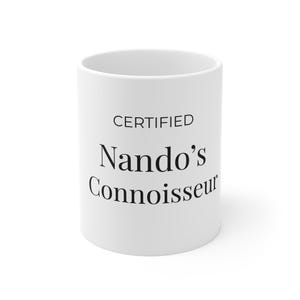 Pode incluir: Uma caneca de cerâmica branca com o texto impresso em preto CERTIFIED Nando's Connoisseur centralizado na parte frontal. A caneca tem um formato cilíndrico com acabamento liso e um design simples e limpo, exibindo o texto numa fonte clara.